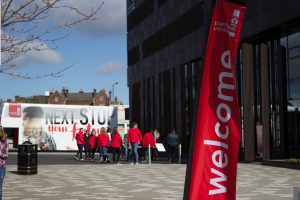 image of entrance to Staffordshire universities Science Center, with student ambassadors and the Staffordshire university tour bus