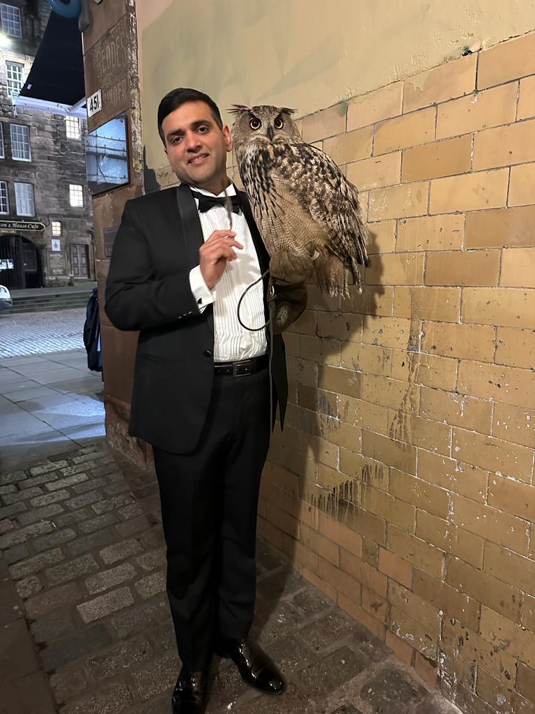 Full length shot of a man with short black hair in a black tuxedo, white shirt and black bow tie holding a large brown owl, standing next to a brick building on a brick paved side street at night