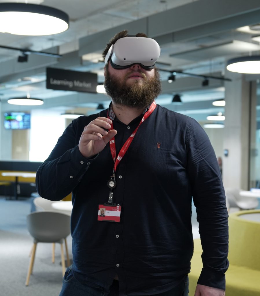 Head and torso shot of a person with a full brown beard wearing a dark blue shirt and a Staffordshire University lanyard. He is standing in the Learning Market in the Catalyst building wearing a virtual reality headset.