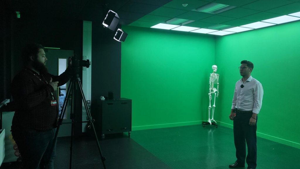 On the left is a person (George) in shadow standing up looking into a camera on a tripod. The camera is pointing towards another person (Edward) standing in a brightly lit half of the room with green walls and flooring. There is a demonstration skeleton hanging in the back corner of this part of the room.