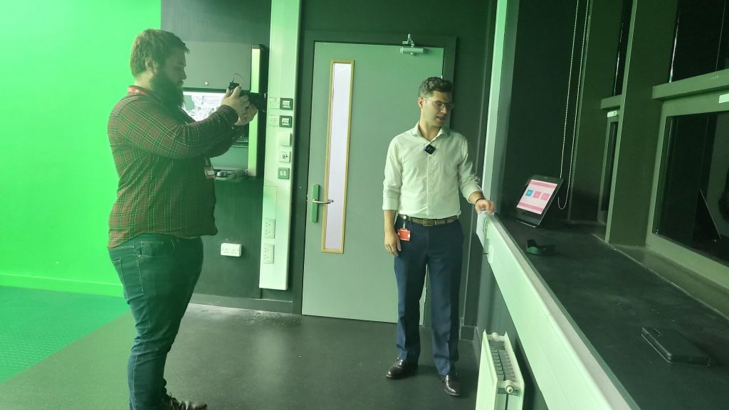 On the left is George standing up pointing a camera towards a tablet screen on top of a deep waist-high window-sill on the right. Edward is standing next to the screen looking towards it, with the door to the room behind him. To the left of the door is a panel of switches and a wall-mounted computer monitor and keyboard (partially obscured)