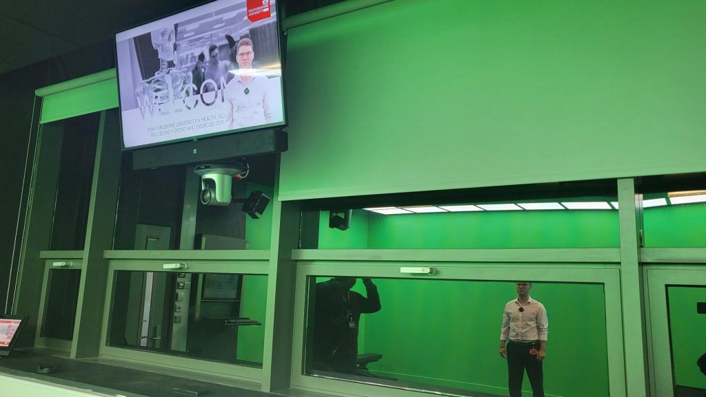 A large monitor screen is shown mounted from the ceiling above a panel of reflective windows, with a camera attached below it. On the screen is a presentation slide, with the image of Edward in the bottom right corner. Edward and George are shown reflected in one of the windows.