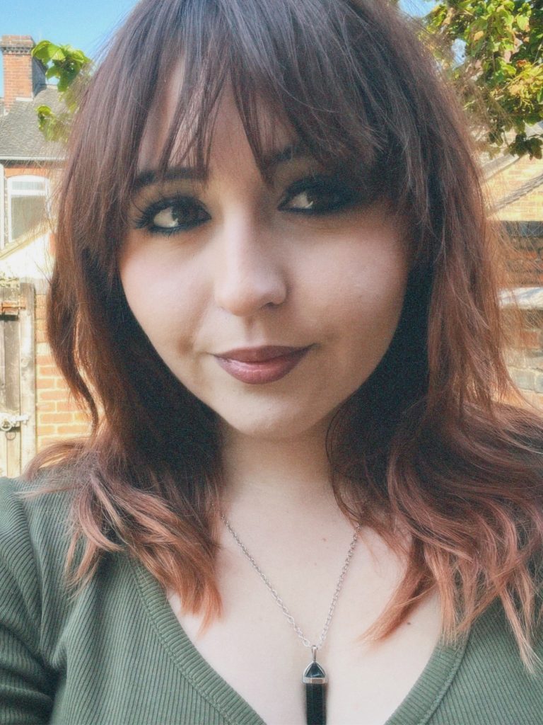 Head and shoulders shot of a person with long brown hair and a fringe, wearing a green top and pendant necklace, standing in front of buildings with a blue sky