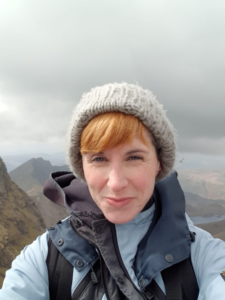 Head and shoulders shot of a person with a light orange fringe under a grey wooly hat wearing a blue walking jacket, with a backdrop of a misty mountain range