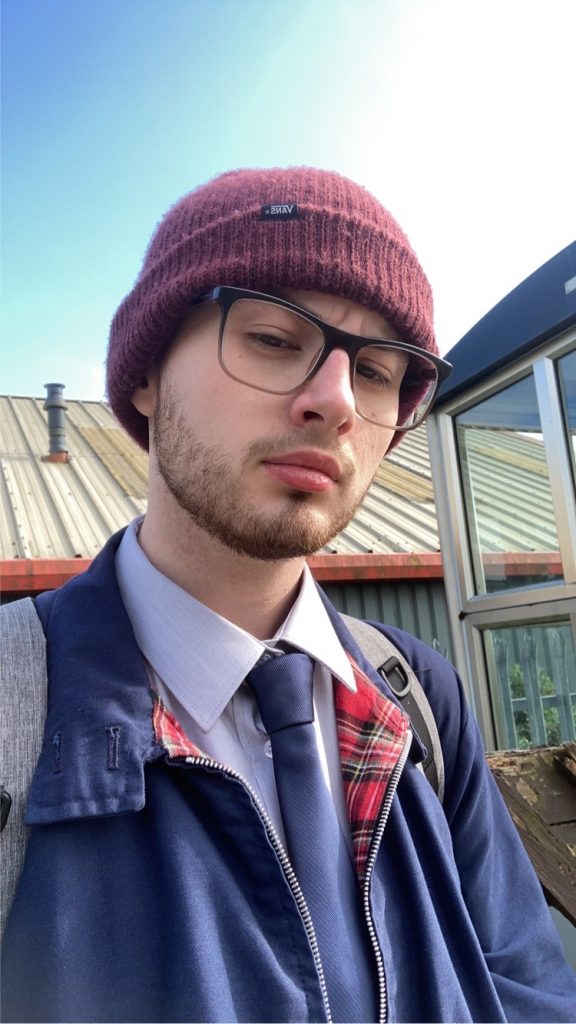 Head and shoulders shot of a person with a short beard and glasses wearing a burgundy hat, blue jacket, shirt and a blue tie, standing outside some buildings with a blue sky