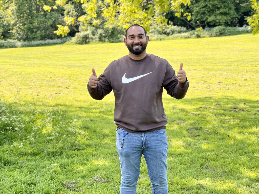 A smiling man with short black hair and beard standing on a grassy lawn with trees in the background, giving a double thumbs up sign. He is wearing a light brown sweatshirt and blue jeans.