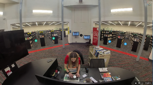 A still image from the Library's thinglink showing a person sat at a desk in the Library Core building surrounded by shelves of books, screens and small, circular, clickable hotspot tags.