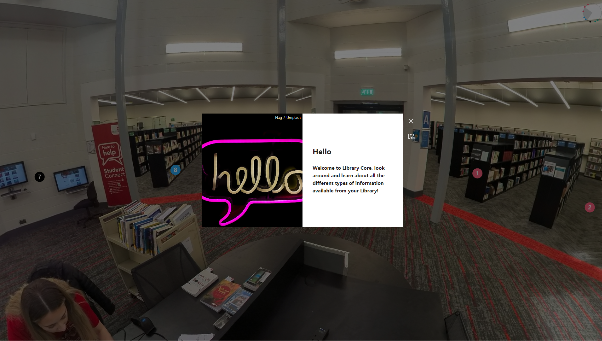 A still image from the Library's thinglink showing a person sat at a desk in the Library Core building surrounded by shelves of books, screens and small, circular, clickable hotspot tags. One of the tags is open in a rectangular window in the centre of the screen with an image reading "hello" on the left and some text on the right.